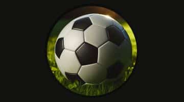 Bilden visar en traditionell fotboll som ligger på en grön gräsmatta. Fotbollen är vit med svarta paneler och ser ut att vara belyst av solljus från vänster sida. Bakgrunden är mörk, vilket framhäver fotbollen och gräset.